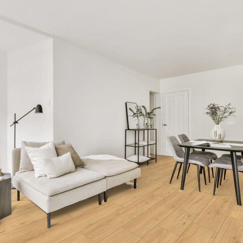 A modern living room featuring a light beige sectional sofa with white and taupe pillows, a black floor lamp, light floating laminate wood flooring, and a dining area with gray chairs and a black table.